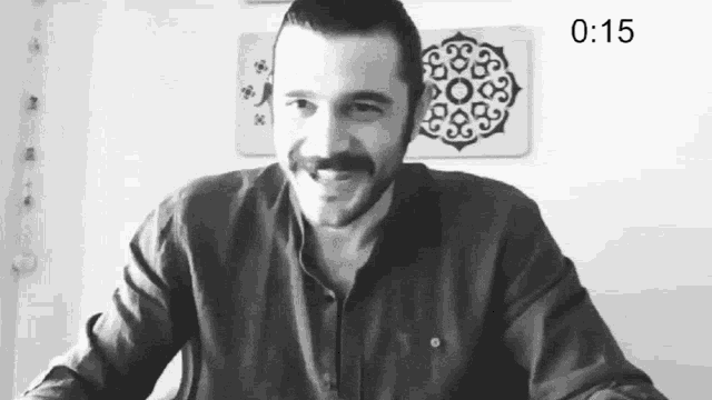 a man with a mustache is smiling in front of a wall with a clock that says 0:15