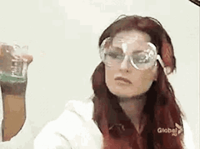 a woman with red hair is wearing goggles and holding a beaker of liquid .