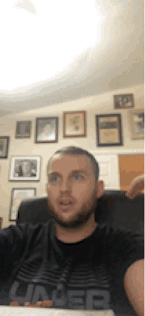a man wearing a black under armour shirt sits in front of a wall of framed pictures