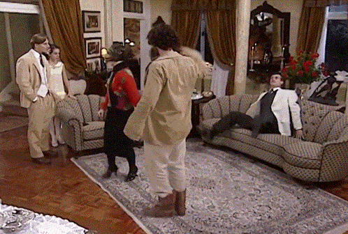 a group of people standing in a living room with a man sitting on a couch