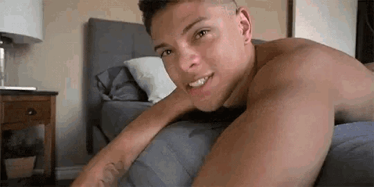a shirtless man is laying on a bed with his head on a pillow and smiling .