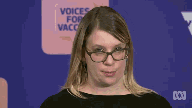 a woman wearing glasses and earrings stands in front of a sign that says voices for vaccines