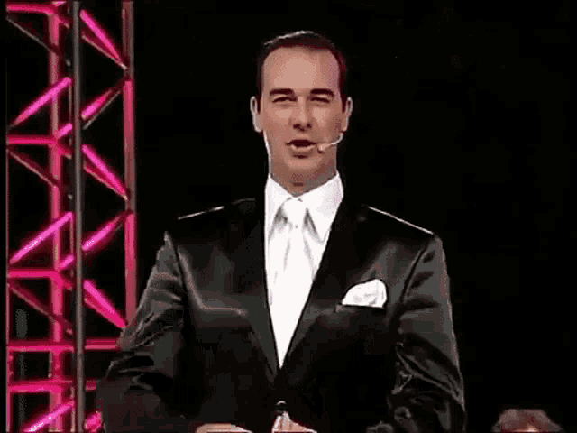 a man in a black suit and white tie is speaking into a microphone .