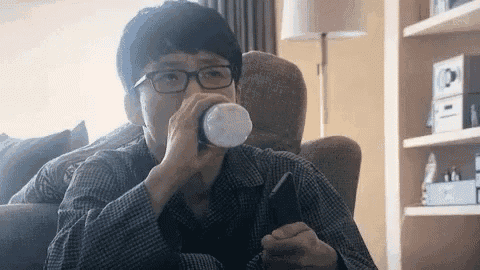 a man wearing glasses is sitting on a couch drinking from a cup while looking at his cell phone .