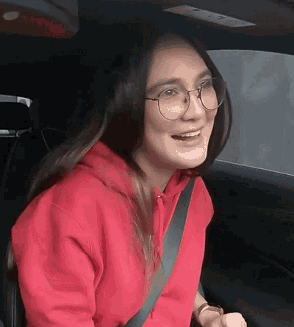 a woman wearing glasses and a red hoodie is smiling while sitting in a car