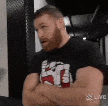 a man with a beard is standing with his arms crossed and wearing a black t-shirt .