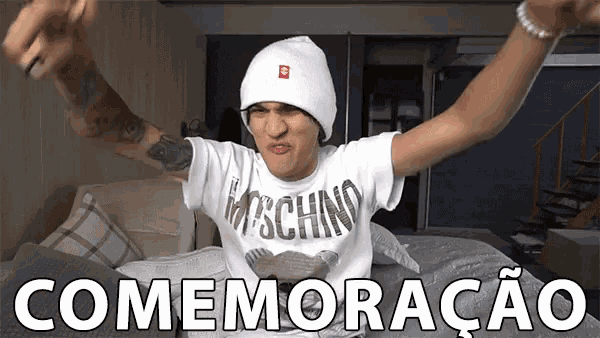 a man wearing a white beanie and a moschino shirt is sitting on a bed with his arms outstretched .