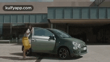 a woman in a yellow skirt is standing next to a green car in front of a building .