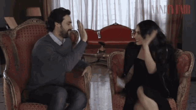 a man and a woman are sitting on a red couch and the word vanity fair is on the curtain