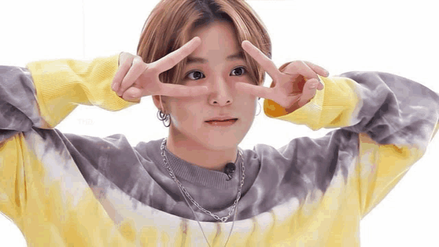 a young man wearing a tie dye sweater and a necklace is making a peace sign with his hands .