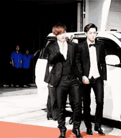 two men in suits are walking on a red carpet in front of a car