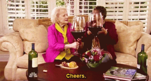 two older women are sitting on a couch drinking wine and toasting .
