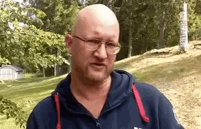 a bald man with glasses and a beard wearing a blue hoodie is standing in a field .