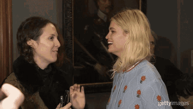 a woman in a blue sweater with flowers on it is talking to another woman in front of a painting