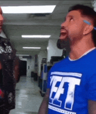 a man wearing a blue shirt that says fft is talking to another man in a hallway .