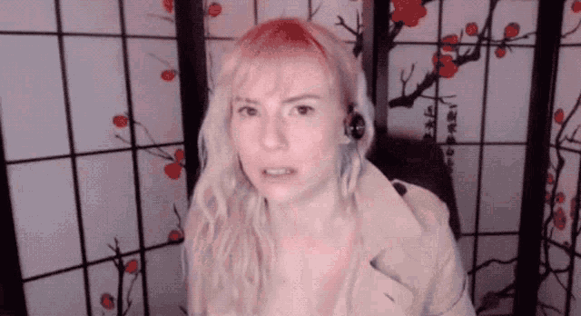 a woman with pink hair is wearing headphones and a trench coat in front of a floral screen .