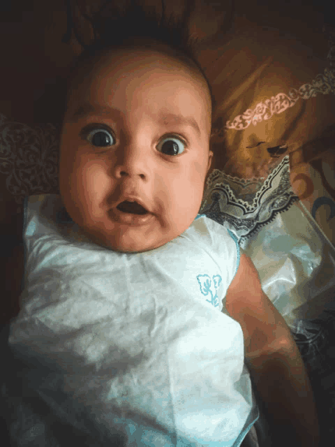 a baby with a surprised look on his face is wearing a white shirt with a blue letter e on it