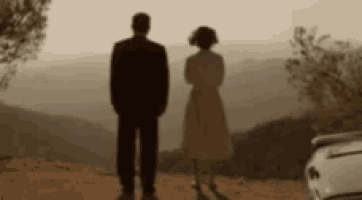 a man and a woman are standing next to each other on top of a hill with a car in the background .