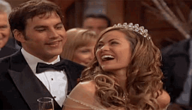 a man in a tuxedo and a woman in a tiara are laughing while holding a glass of champagne .