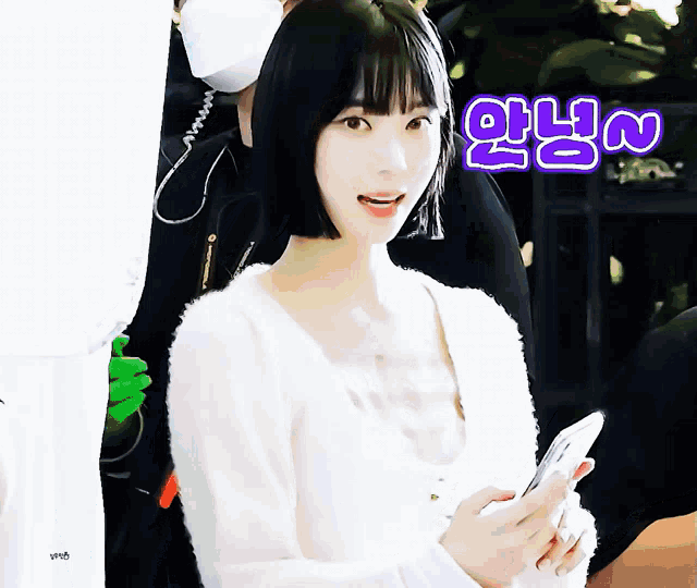 a woman wearing a white sweater holds a cell phone in front of a sign that says ' 안녕 ' on it