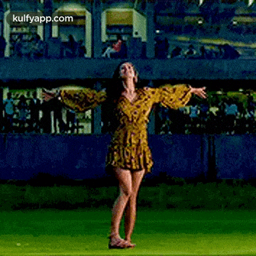 a woman in a yellow dress is standing on top of a lush green field with her arms outstretched .