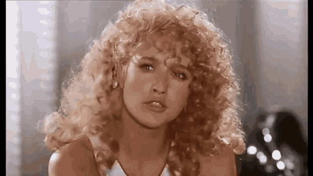 a woman with curly blonde hair and bangs is looking at the camera .