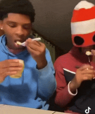 a boy in a blue hoodie eating a sandwich next to another boy in a red and white hat