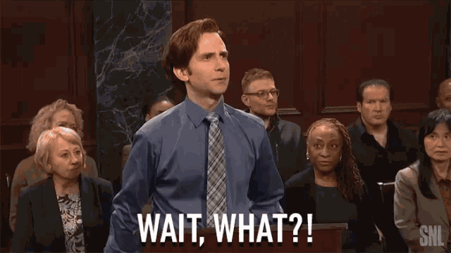 a man in a blue shirt and tie stands in front of a group of people and says " wait what "