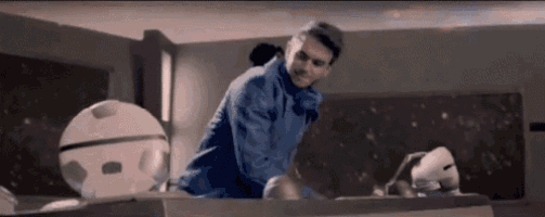 a man in a blue jacket is standing on a table in a room with a robot .