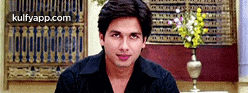 a man in a black shirt is sitting in front of a vase of flowers and looking at the camera .