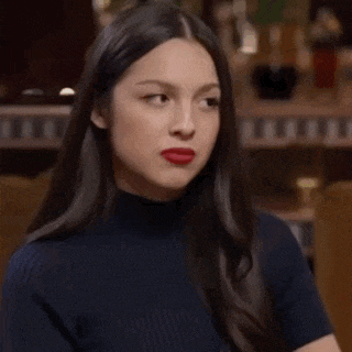 a woman with long hair and red lipstick is wearing a black turtleneck and making a funny face .