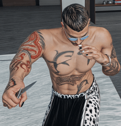 a shirtless man with tattoos on his arms and chest holds a knife