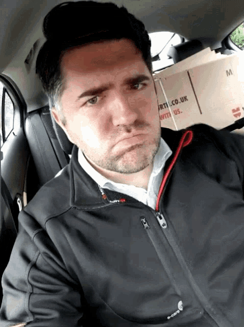 a man in a black jacket is sitting in a car with a cardboard box that says art.co.uk