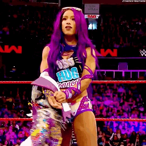 a woman with purple hair is standing in a wrestling ring wearing a shirt that says ' hug ' on it .