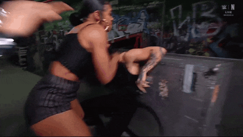 two women are fighting each other in front of a wall with graffiti on it .