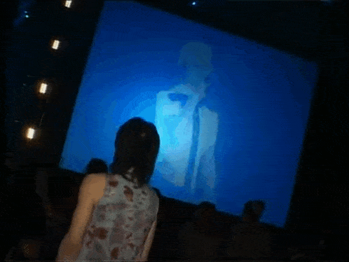 a woman stands in front of a large screen that shows a man singing