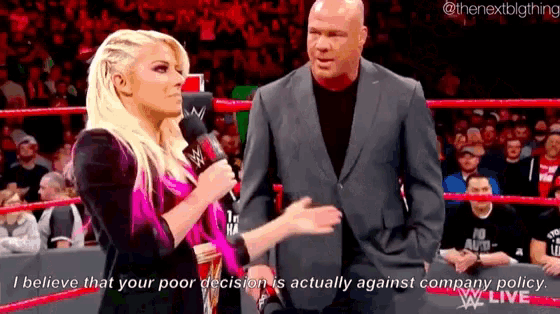 a woman is talking into a microphone in a wrestling ring while a man in a suit stands behind her .
