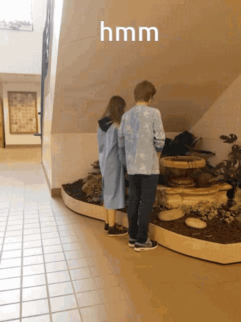a boy and a girl are looking at a fountain in a hallway with the word hmm above them