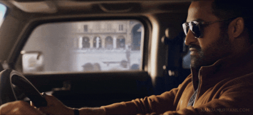 a man wearing sunglasses is driving a car with nandamurifans.com written on the bottom of the screen