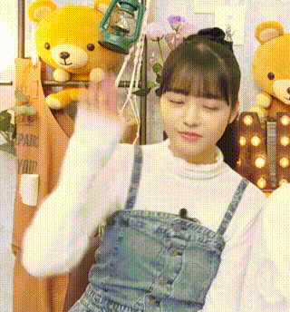 a girl wearing overalls and a white sweater is waving her hand in front of a stuffed teddy bear .