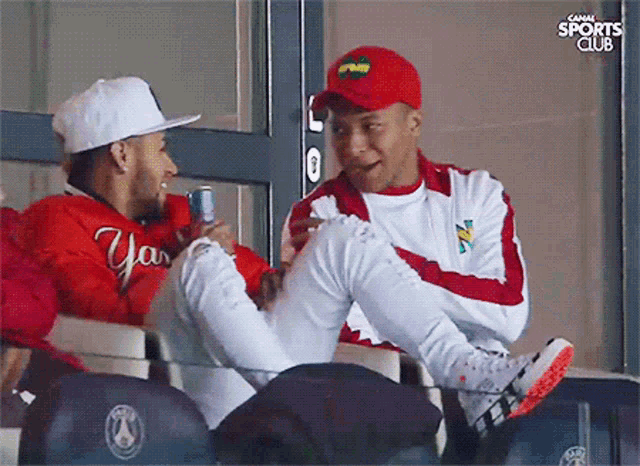 a man in a red jacket with the word yas on it sits next to another man in a white jacket
