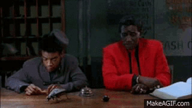 two men are sitting at a table in a room talking to each other . one of the men is wearing a red jacket .