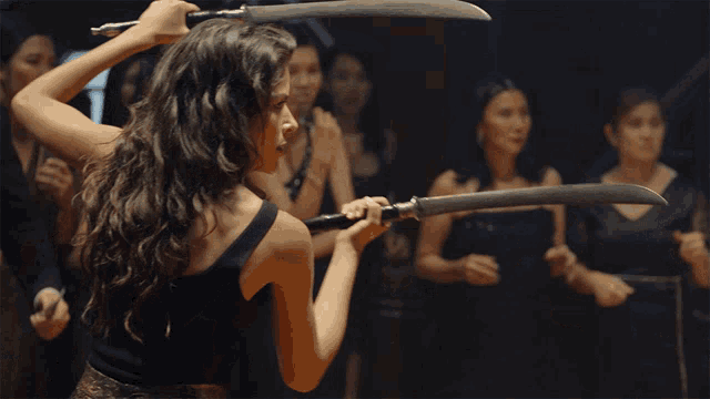 a woman in a black tank top is holding two swords in front of a group of women