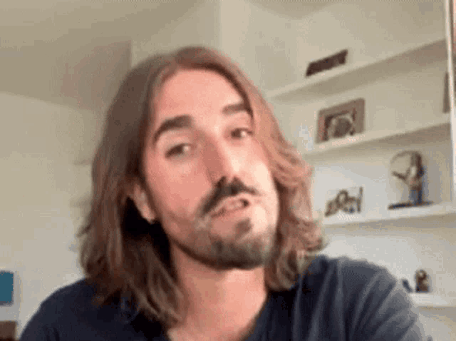 a man with long hair and a beard is sitting in front of a shelf .