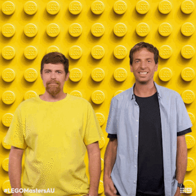 two men are standing in front of a wall of yellow lego bricks
