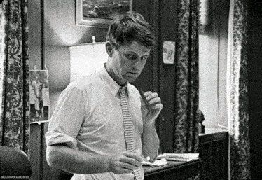 a black and white photo of a man in a white shirt and tie standing in a room .