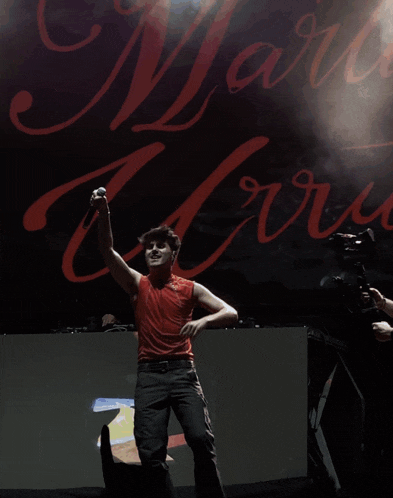 a man in a red tank top stands on stage in front of a sign that says " mario "