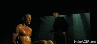 a man without a shirt is sitting in a chair in a dark room while a man in a suit stands .