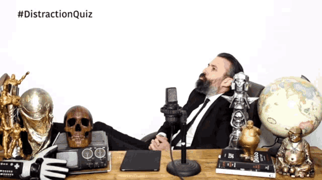a man in a suit sits at a desk with a microphone in front of a globe and a skull