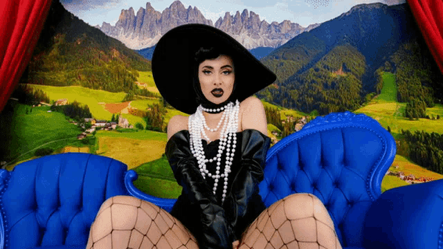 a woman in a black hat and pearls sits on a blue couch with mountains in the background
